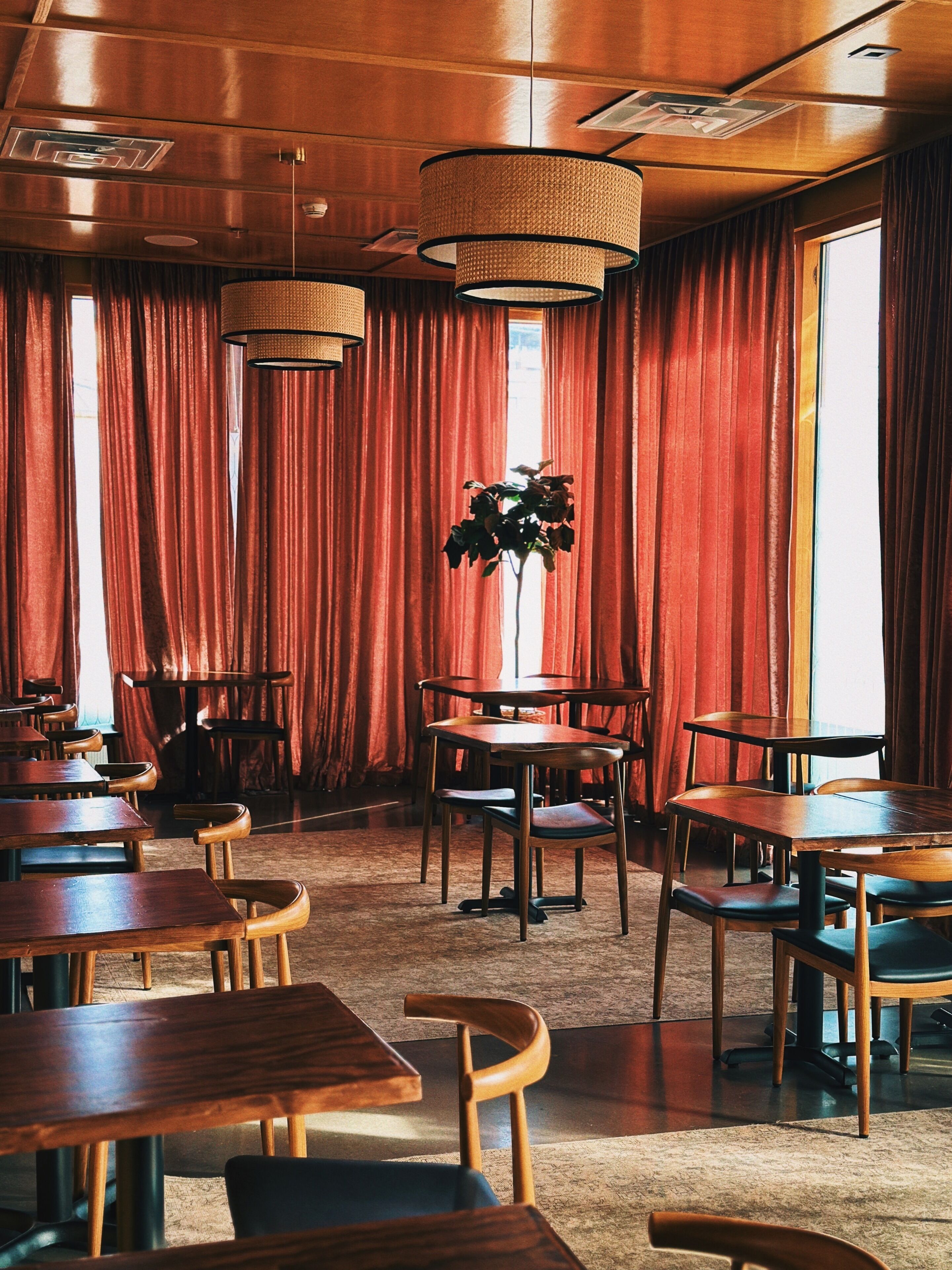 Un ristorante con tavoli e sedie di legno, tende rosse e lampade a sospensione.
