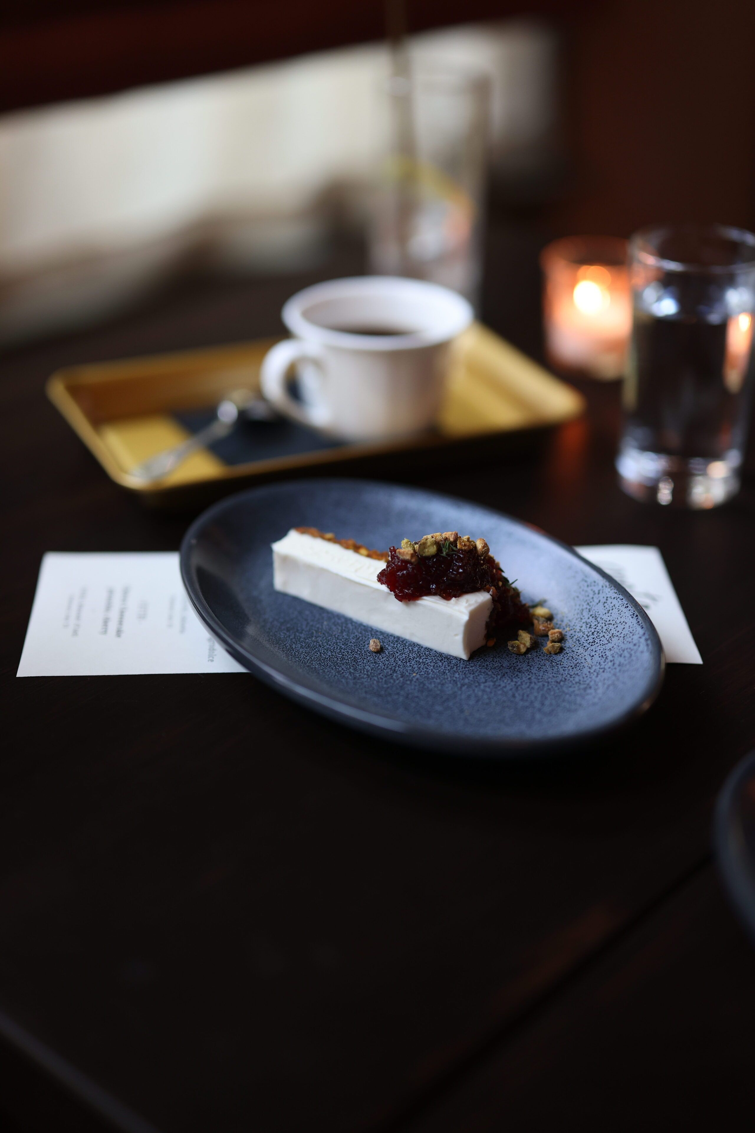 A dessert plate with a piece of cheesecake and fruit on a dark blue plate, a cup of coffee, a candle, and a glass of water on a table.