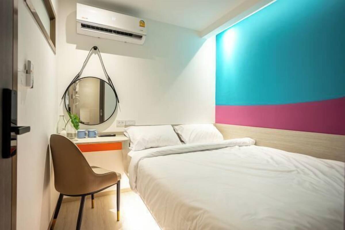 A hotel room with a bed, a desk with a chair, a round mirror, and a wall with a blue and pink design.