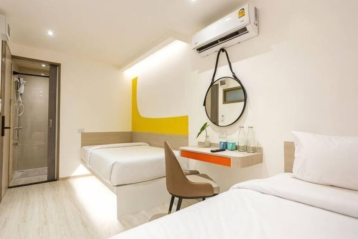 A hotel room with a bed, a desk, a chair, a mirror, and an air conditioning unit.