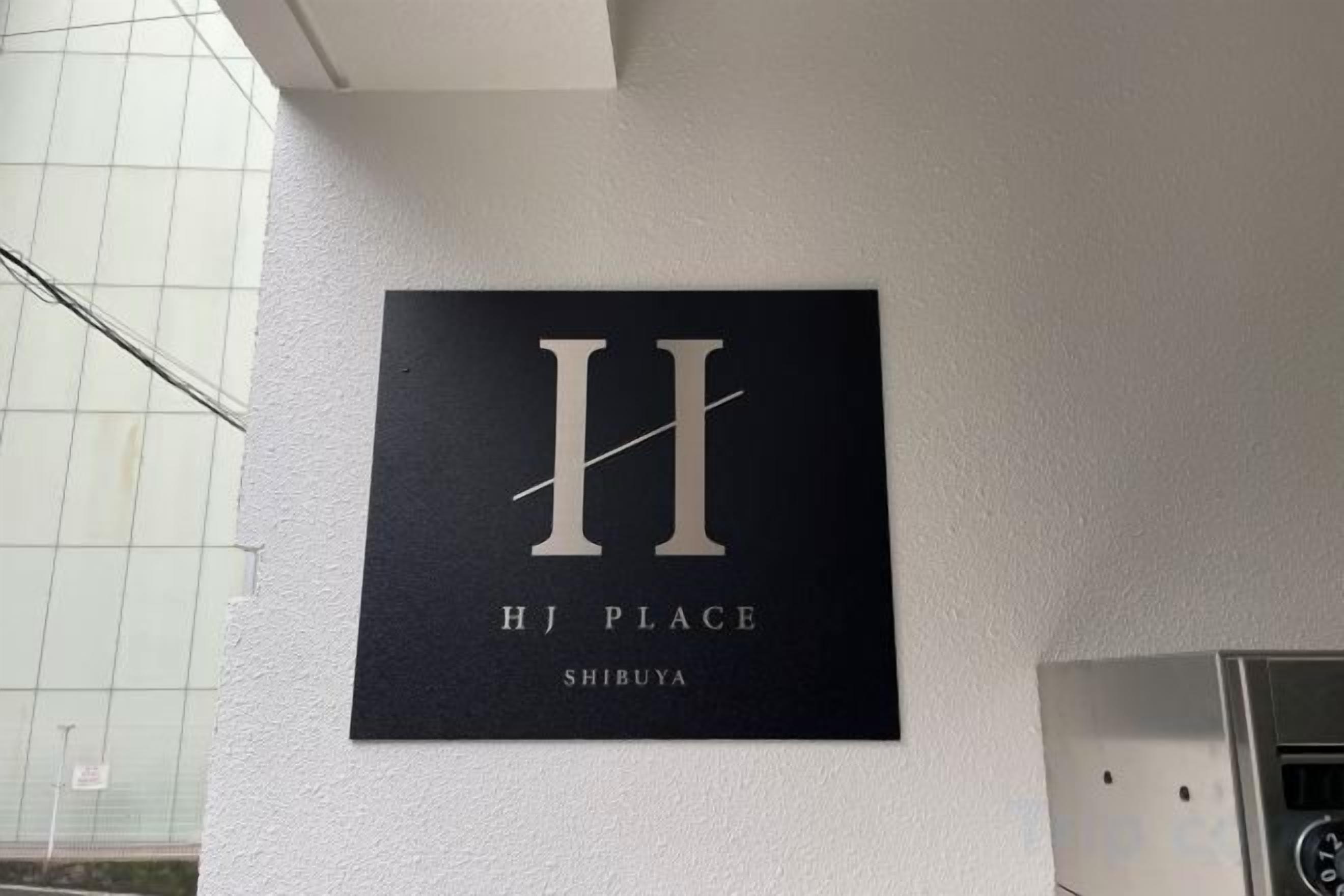 A sign with the letter 'H' and the words 'HJ PLACE SHIBUYA' on a white wall.
