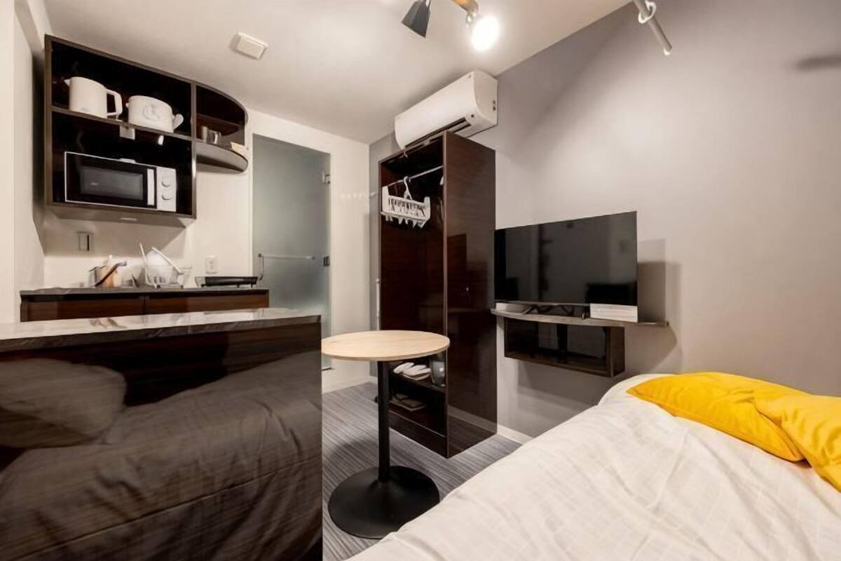 A modern hotel room with a bed, a small round table, a TV, a wardrobe, and a kitchenette area.
