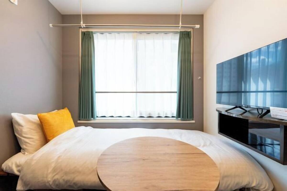 A hotel room with a bed, a round wooden table, a TV mounted on the wall, and a window with curtains.