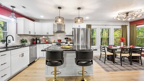 A modern kitchen with white cabinets, a central island, two black bar stools, and a dining area with a table and chairs. - Lodge-style Home Near Gatlinburg w/ Theater, Hot Tub & Game Room Evergreen Escape by Avantstay (Gatlinburg)