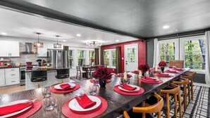 A modern kitchen with white cabinets, stainless steel appliances, and a dining area set for a meal with red tableware and decor. - Lodge-style Home Near Gatlinburg w/ Theater, Hot Tub & Game Room Evergreen Escape by Avantstay (Gatlinburg)