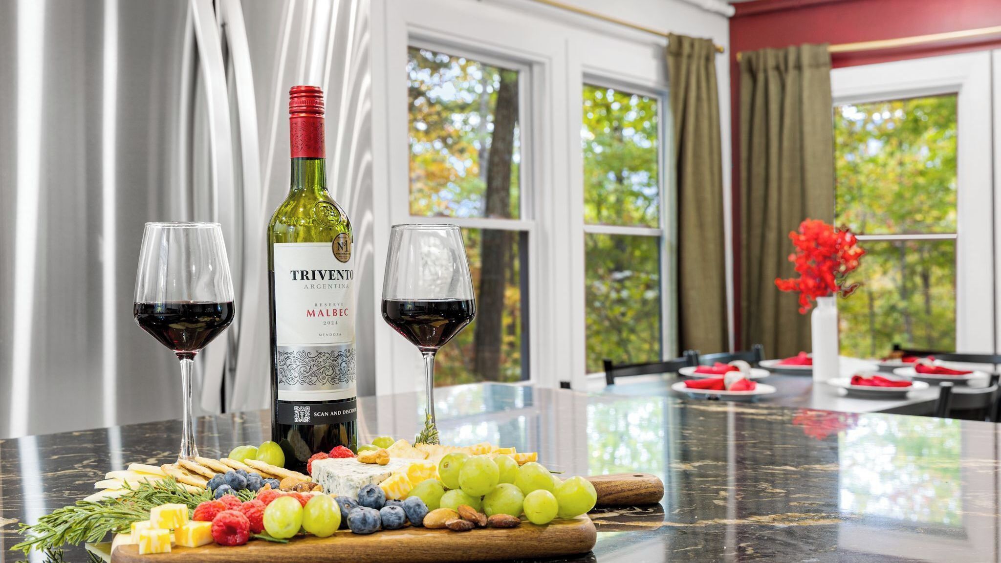 A table with a bottle of wine, two glasses, and a cheese board with fruits and nuts.