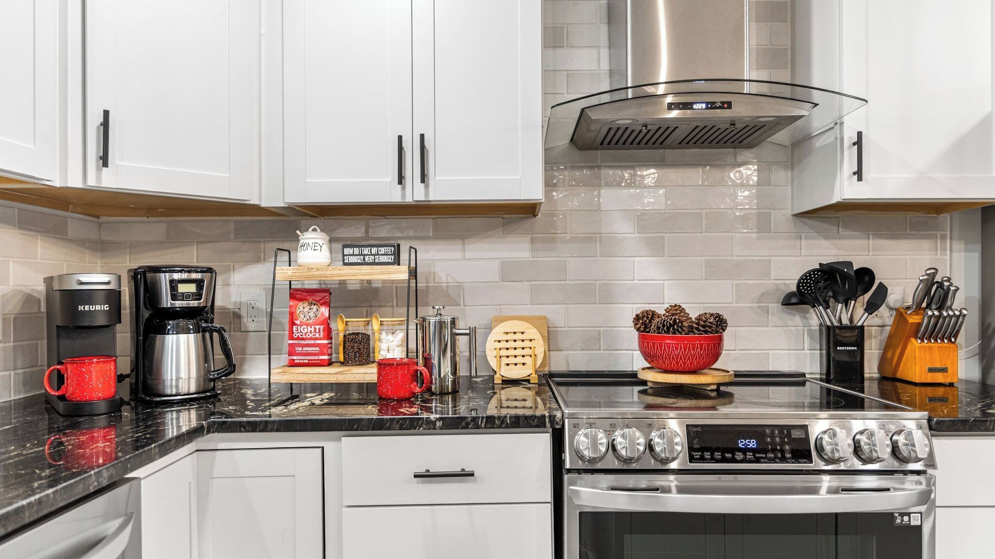 A modern kitchen with white cabinets, a stainless steel range, a coffee maker, a red mug, a coffee grinder, and a bowl of pinecones.