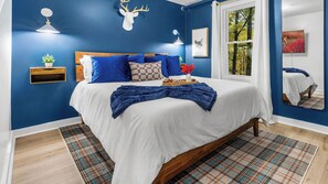 A bedroom with a large bed, a wooden headboard, a mounted deer head, a bedside table with a plant, and a window with a view of trees.