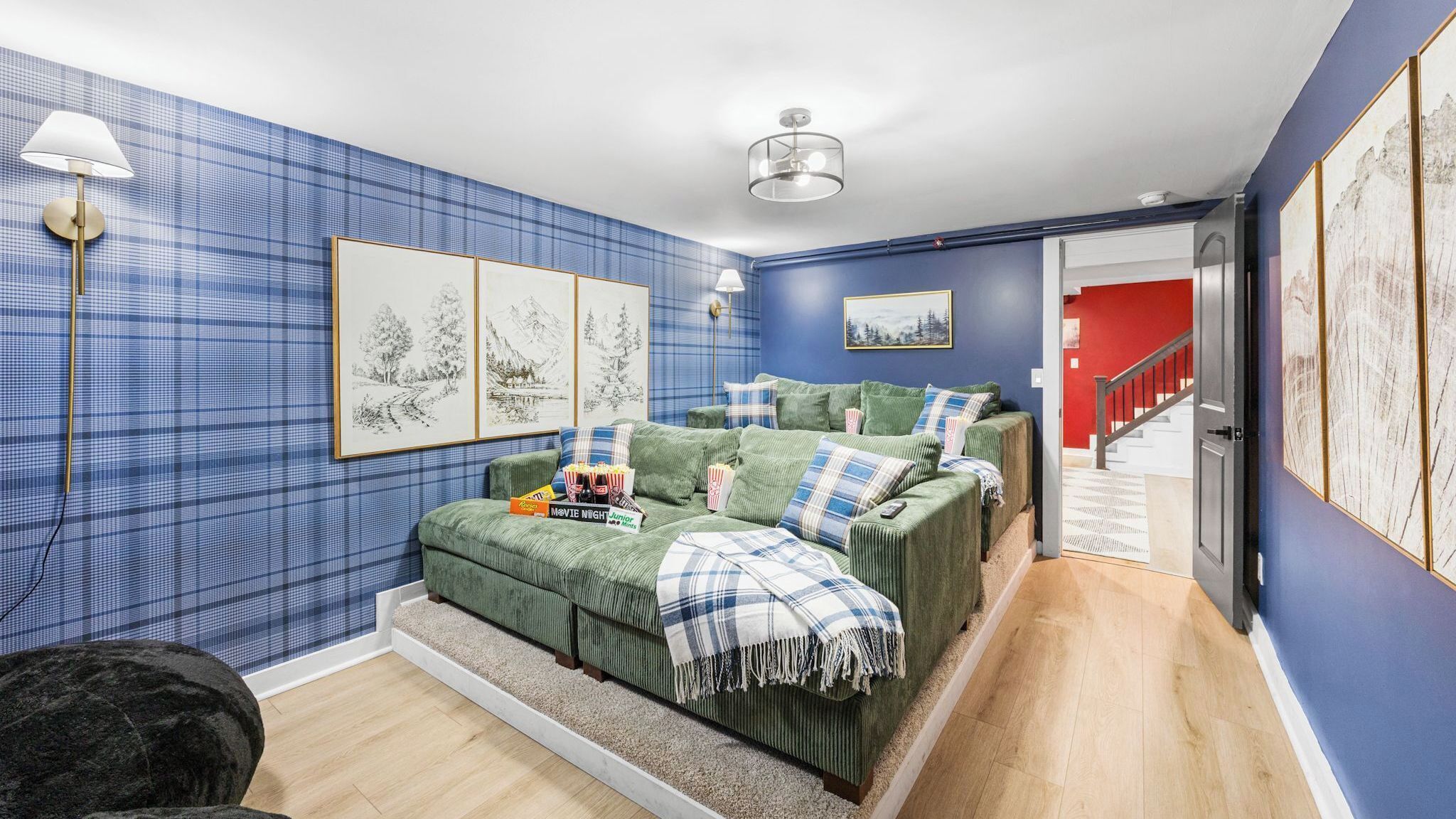 A living room with a green sofa set, framed artwork on the walls, and a staircase leading to a red room.