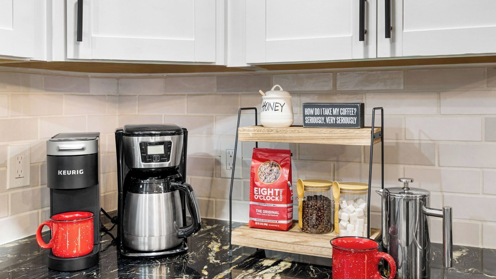 A kitchen with a Keurig coffee maker, a coffee pot, a mug, a sign, a bag of Eight O'Clock coffee, a honey jar, and a sugar container.