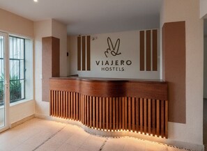 A reception desk with a logo for Viajero Hostels, a wooden counter with vertical slats, and a window to the left.