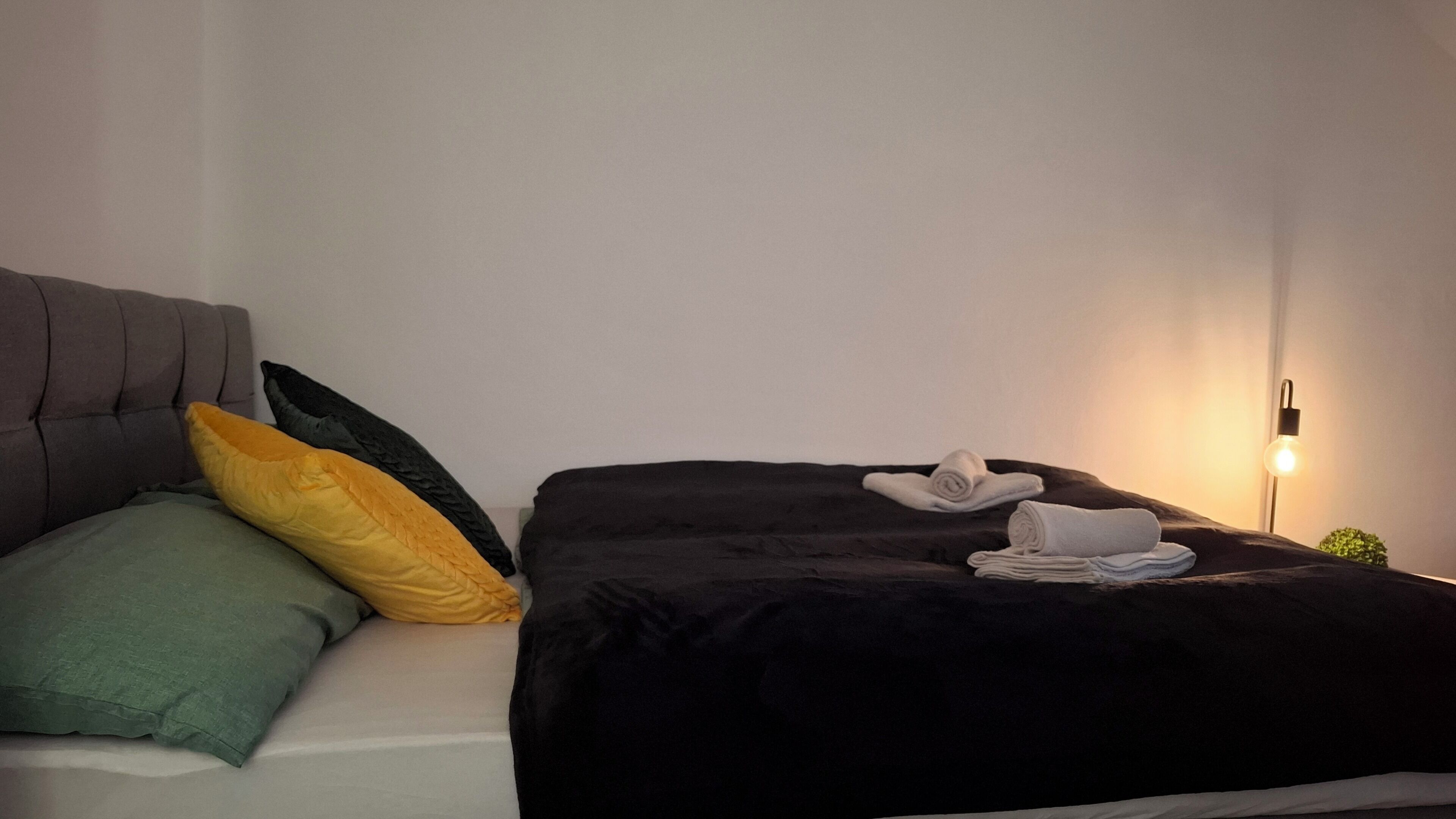 A neatly made bed with a dark blanket and pillows, a folded towel, and a bedside lamp.