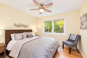 A bedroom with a bed, nightstands, a chair, a ceiling fan, and a window with a view of trees.