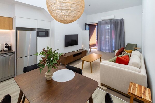 A modern living room with a sofa, coffee table, and dining table. There is a TV mounted on the wall and a kitchen area with a refrigerator and microwave.