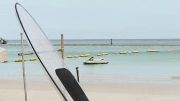 A beach with a surfboard leaning against a post, a small boat in the water, and a wooden structure in the background.
