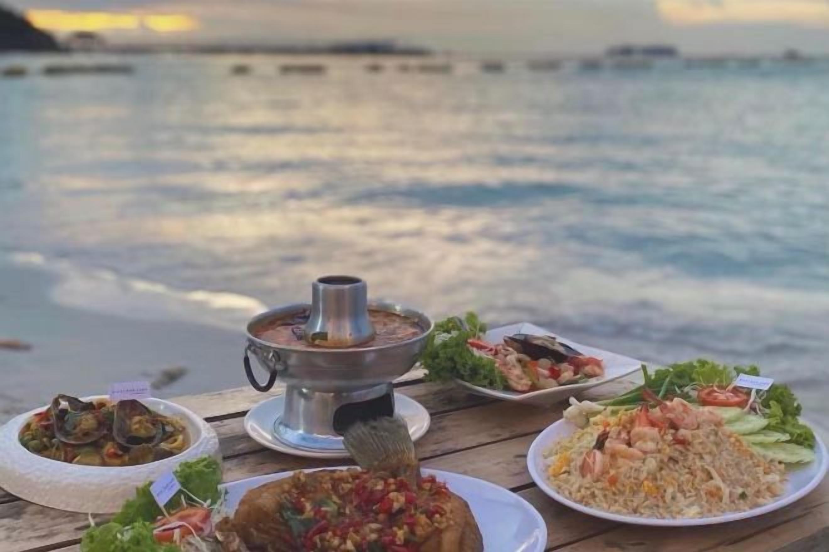 A table with various dishes and a hot pot, set against a backdrop of a calm sea and distant shoreline.