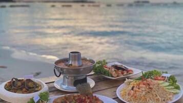 A table with various dishes and a hot pot, set against a backdrop of a calm sea and distant shoreline.