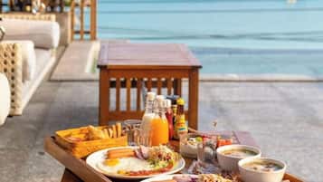 A breakfast tray with eggs, sausages, and a drink, set on a table by a poolside with a view of the ocean.