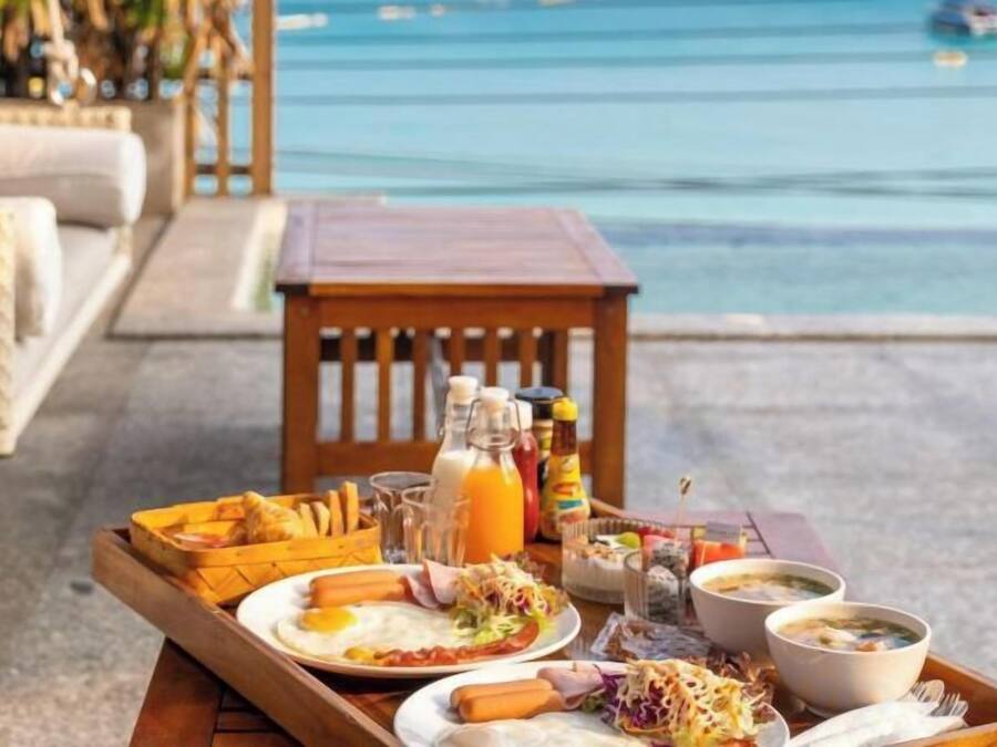 Bandeja de desayuno con huevos, salchichas y una bebida, colocada sobre una mesa junto a una piscina con vista al océano.