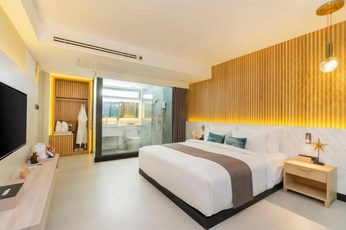 A modern hotel room with a large bed, a bedside table, a TV, and a bathroom visible through an open door.