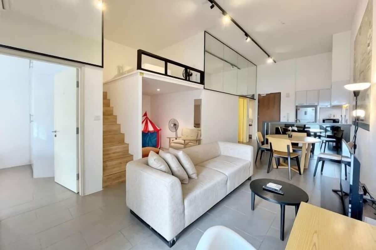 Una sala de estar moderna con sofá, mesa de comedor y sillas, una escalera que conduce a un nivel superior y una zona de cocina.