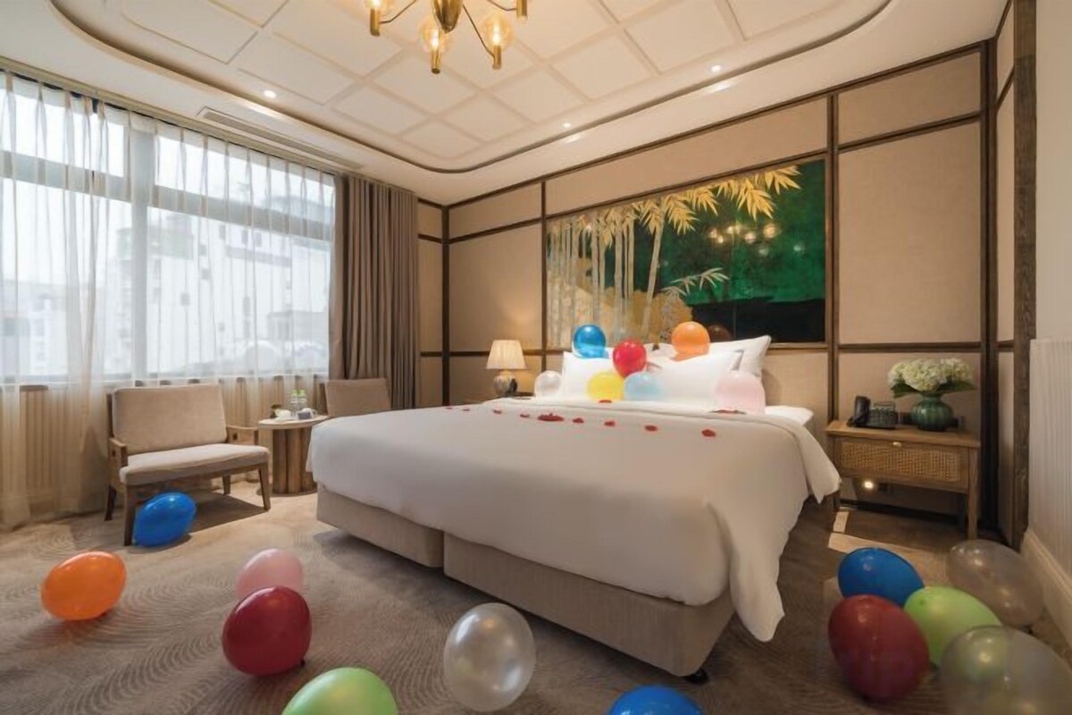 A hotel room with a large bed, a seating area, a nightstand, and a view of the city.