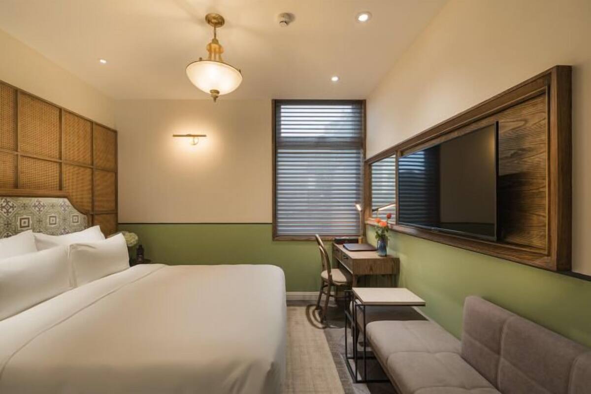 Ein Hotelzimmer mit Bett, Schreibtisch, Stuhl, Sofa, einem großen Spiegel und einem Fenster mit Jalousien.