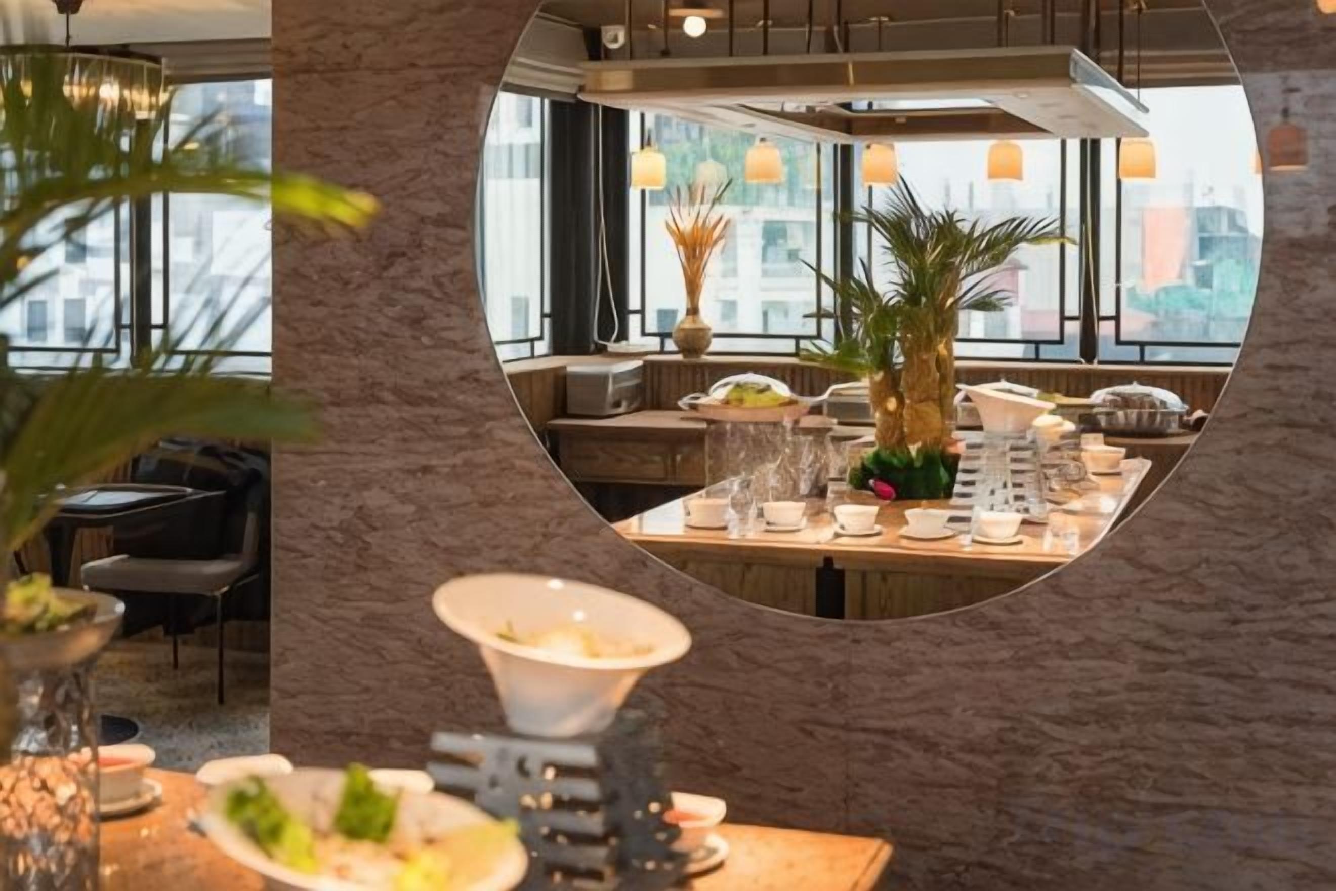 Een restaurant met een ronde spiegel die het interieur weerkaatst, een gedekte eettafel met borden en glazen, en een potplant.