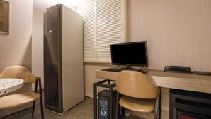 Une pièce comprenant un bureau, deux chaises, un écran d’ordinateur, un clavier, une petite table, un réfrigérateur et une fenêtre avec des stores.