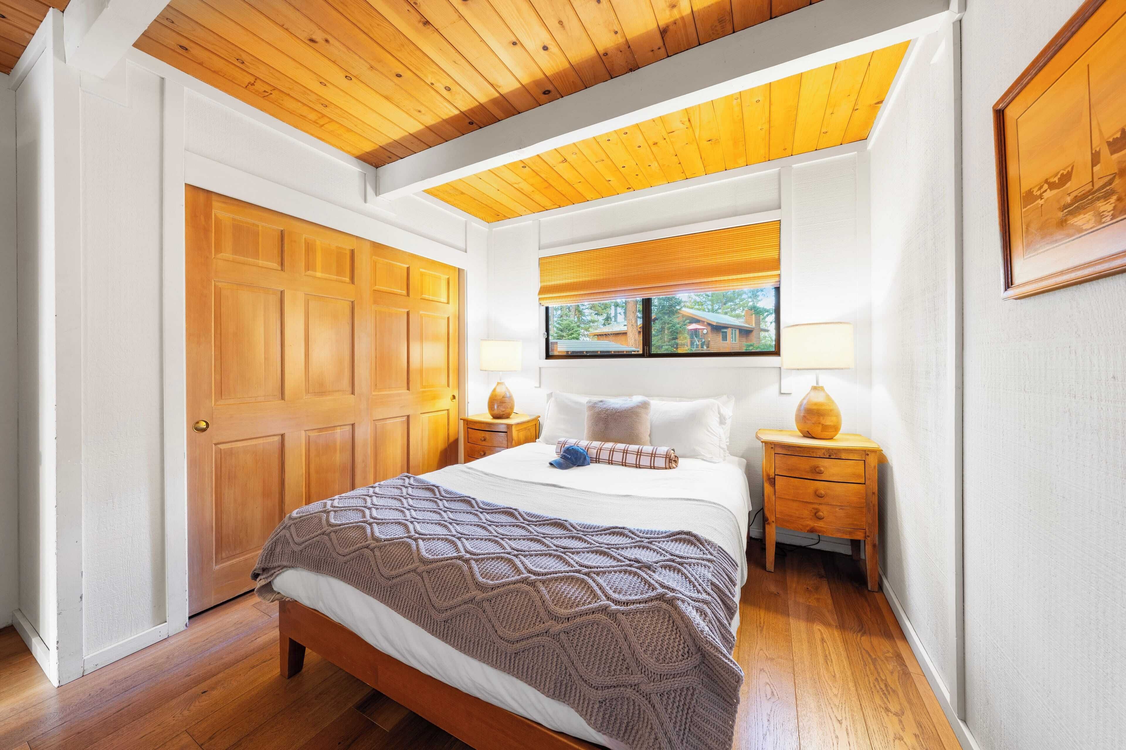 Une chambre avec un plafond en bois, un grand lit recouvert d’une couverture à motifs, deux tables de chevet avec des lampes et une fenêtre avec des stores.