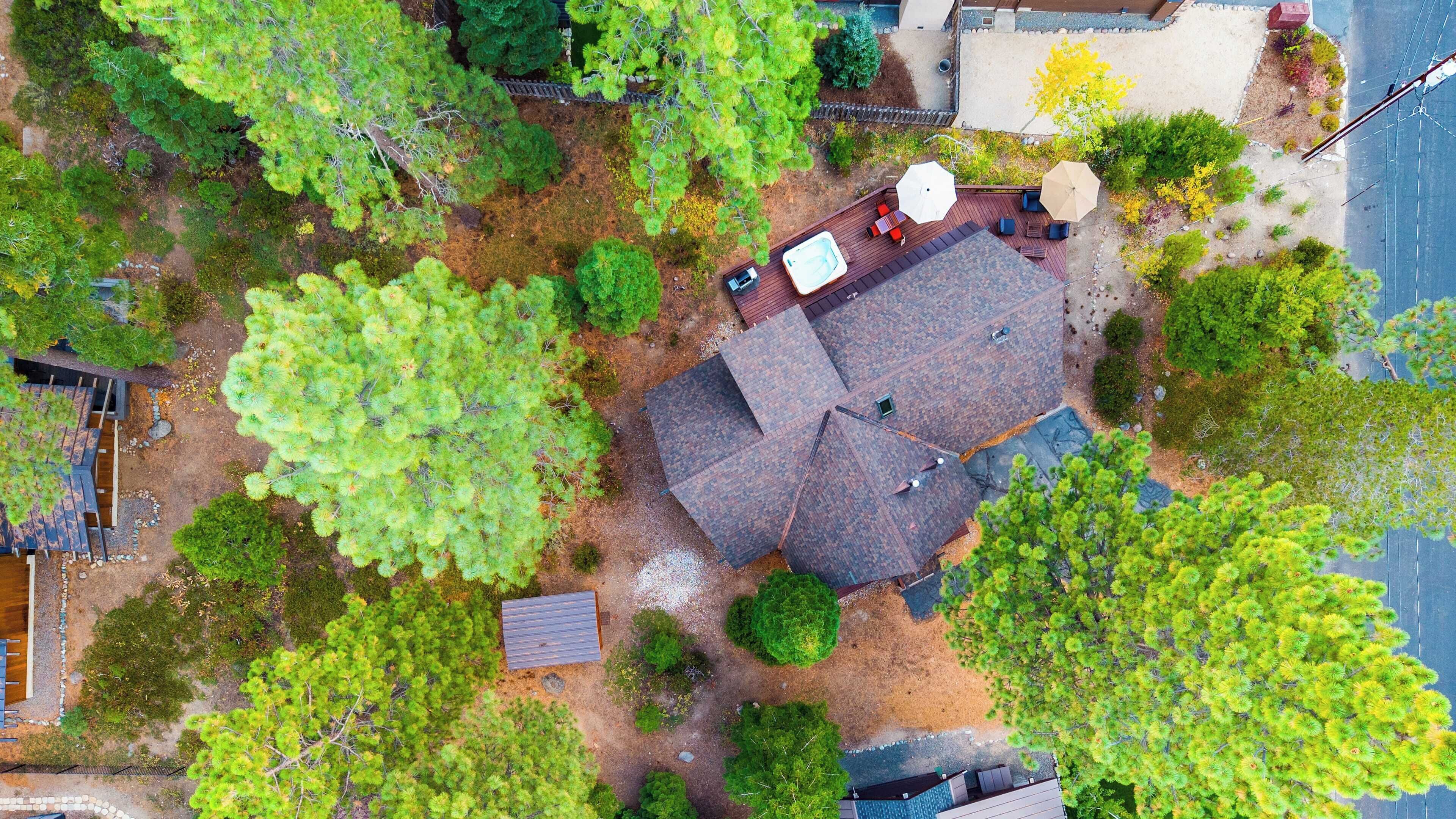 An aerial view of a residential area with a house featuring a sloped roof, a deck with outdoor furniture, and surrounding greenery.