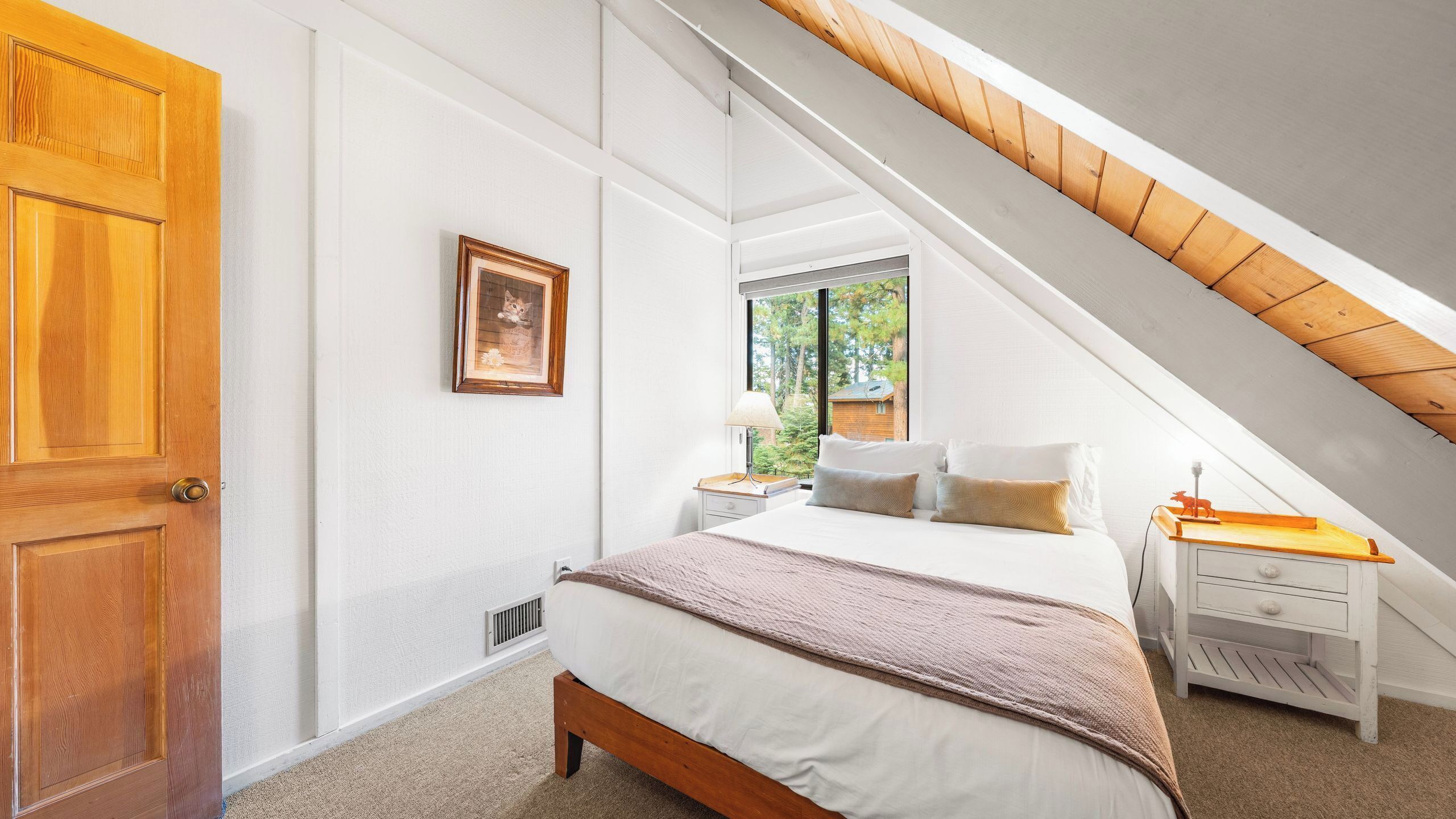 Une chambre avec un plafond en pente, une porte en bois, une table de chevet avec une lampe, un lit avec des oreillers et une fenêtre donnant sur des arbres.