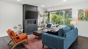 A modern living room with a dark wood floor, a blue sofa, an orange armchair, a wooden coffee table, and a flat-screen TV mounted on a dark wall. - Modern Urban Retreat, Game Room (Portland)
