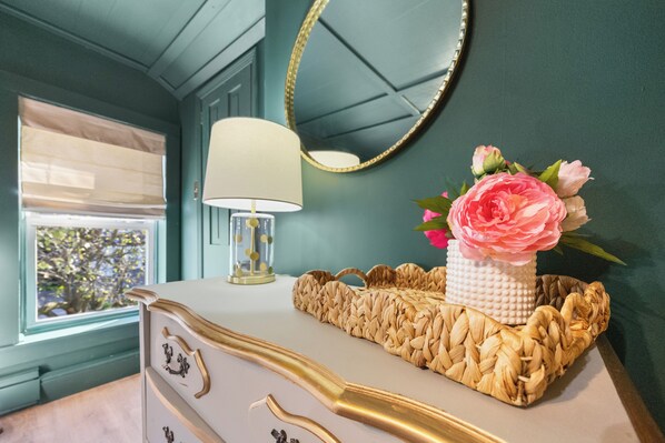 A room with teal walls, a white dresser with gold handles, a woven basket on the dresser, a white lamp, and a mirror with a gold frame.