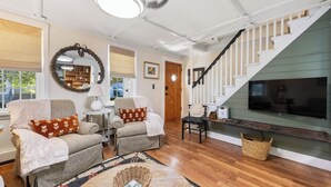 A cozy living room with two armchairs, a coffee table, and a television. A staircase with a wooden railing leads to another level. - Walk Downtown! SUNY, near Power Plants, Hospital, Pets. 3 bathrooms, King beds! (Oswego)