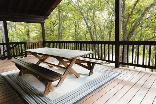 A wooden deck with a picnic table and benches, surrounded by a railing, overlooking a forested area.