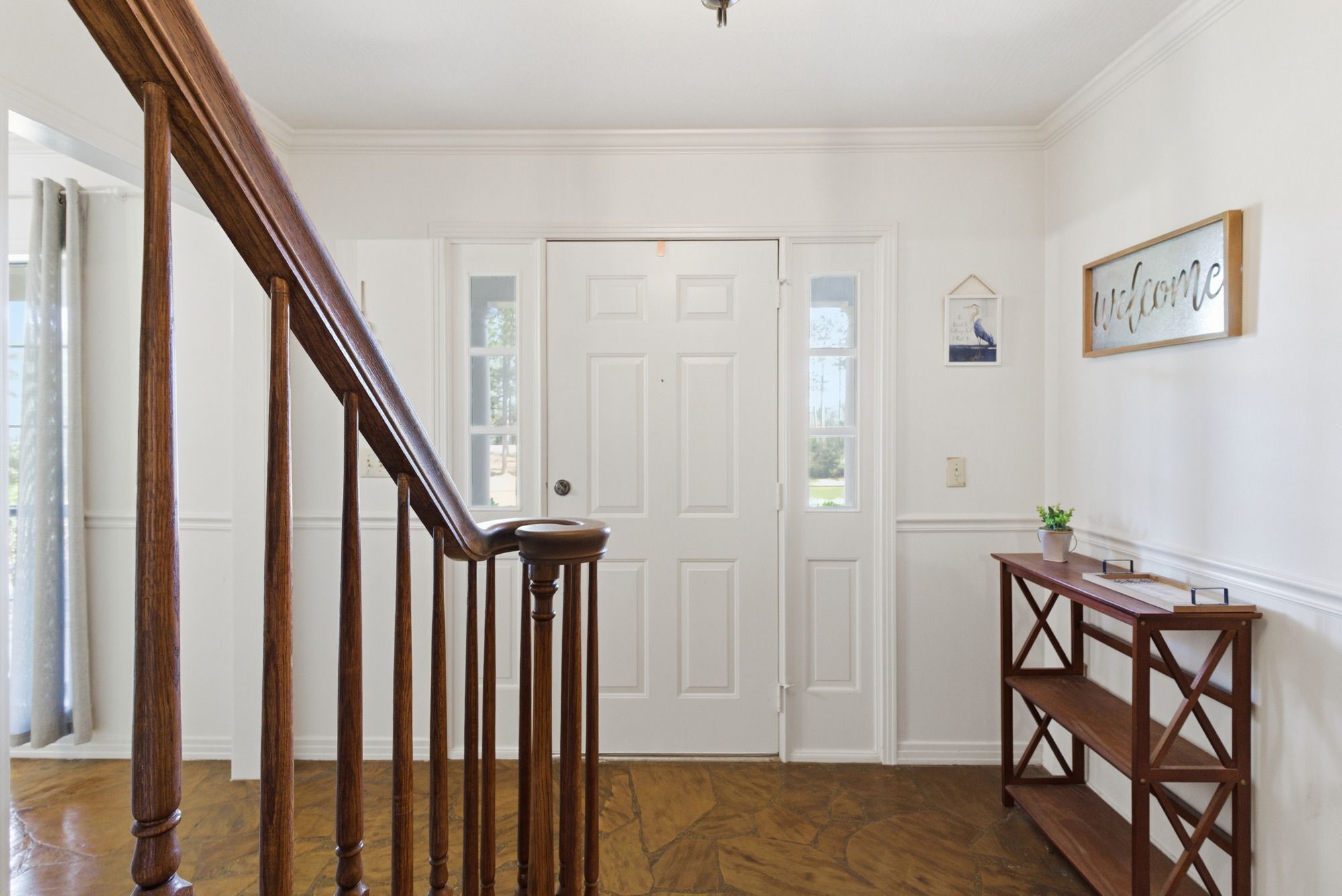 Un escalier en bois menant à une porte blanche, une petite console en bois ornée d’une plante et un panneau d’accueil fixé au mur.