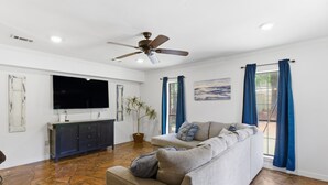 Un salon doté d’un sol imitant le motif de la pierre, d’un canapé gris, d’un ventilateur de plafond, d’un téléviseur à écran plat et d’une fenêtre ornée de rideaux bleus.