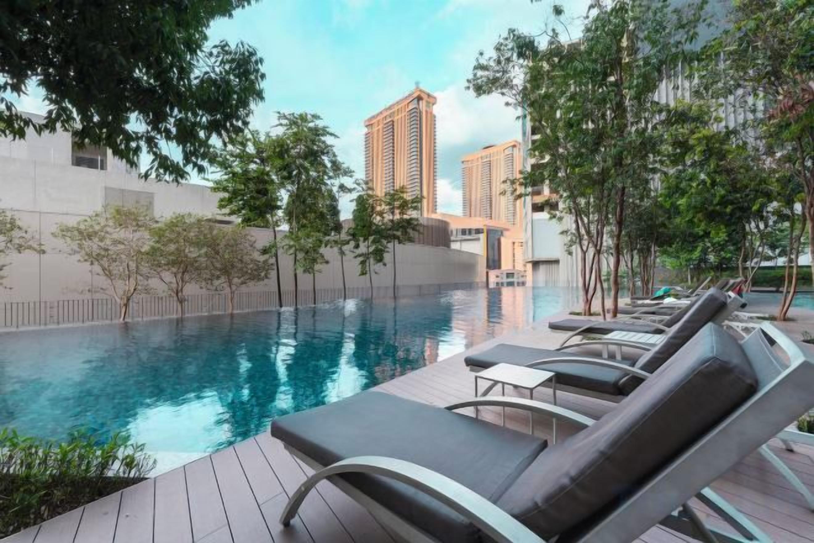 Une piscine entourée d’arbres et de bâtiments, avec des chaises longues à proximité.