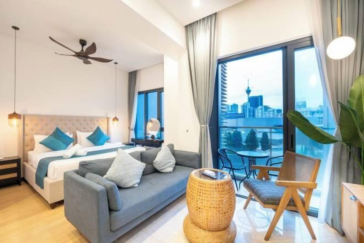 Ein modernes Hotelzimmer mit Bett, Sofa, Esstisch und Stühlen, einem Balkon mit Blick auf die Stadt und einem Deckenventilator.