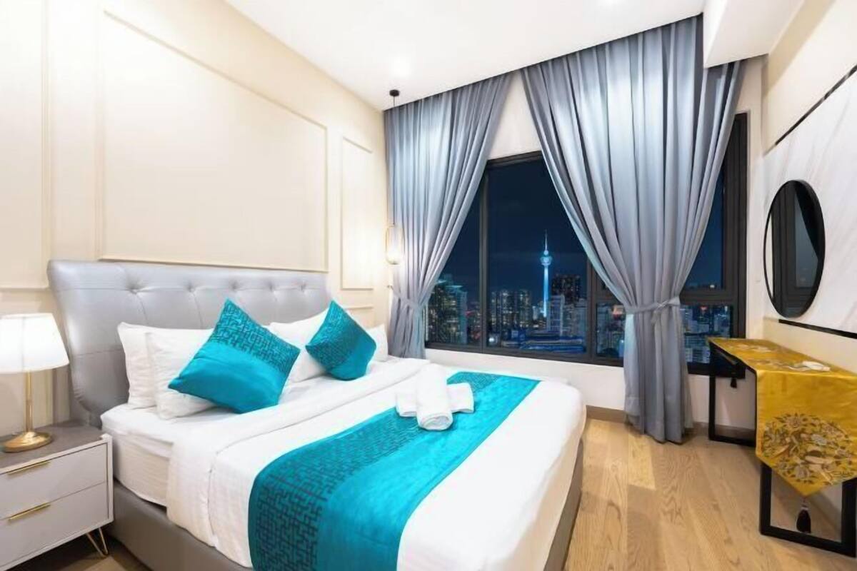 Ein Hotelzimmer mit einem großen Bett, einem Nachttisch, einem Tisch mit goldener Tischdecke und Blick auf eine nächtliche Skyline.