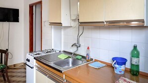 Cocina con estufa a gas, lavamanos y alacenas. Hay una silla y una mesa con mantel.