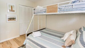 Une chambre avec un lit superposé, une couvre-lit à rayures, une serviette blanche et des oreillers. Le lit supérieur est surmonté d’un panneau en bois, et la pièce comprend un placard aux portes à persiennes.