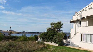 Un bâtiment moderne de couleur blanche, doté d’un escalier en colimaçon, offrant une vue sur la mer et comprenant un espace verdoyant avec des arbres.