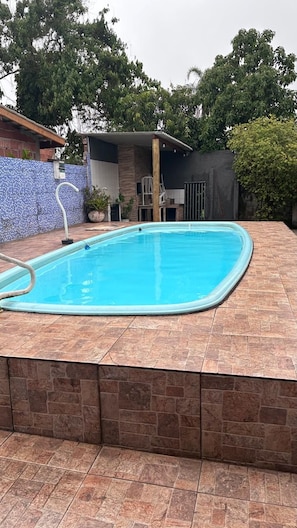 A rectangular swimming pool with a tiled deck, surrounded by a stone wall and a metal fence. - Townhouse with pool and air conditioning near Caraguatatuba shopping mall (Caraguatatuba)
