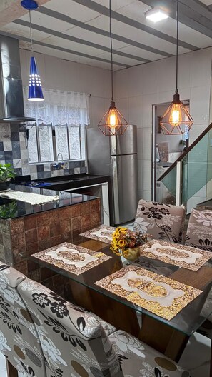 A modern kitchen with a glass dining table, patterned cushions, and pendant lighting. - Townhouse with pool and air conditioning near Caraguatatuba shopping mall (Caraguatatuba)