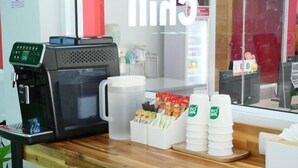 A coffee machine, a coffee cup, and a stack of disposable cups on a wooden counter.