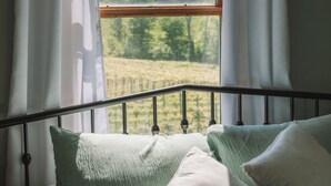 Ein Bett mit Kissen und Blick auf ein grünes Feld durch ein Fenster mit Vorhängen.