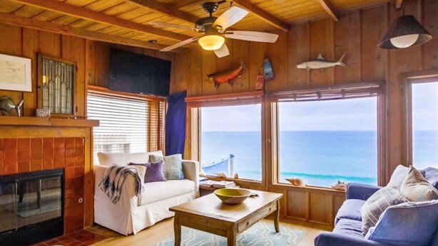 Una habitación acogedora con chimenea, ventilador de techo y ventana grande con vista al océano.