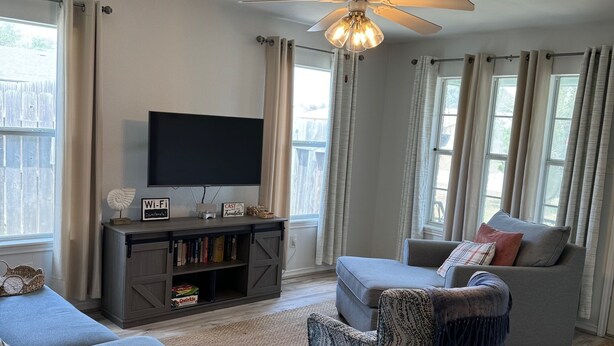 A living room with a ceiling fan, a flat-screen TV, a gray sofa, a patterned armchair, and a bookshelf with books and a Wi-Fi sign. - Hook, Relax & Stay: 3 BR Fishing Haven in Aransas Pass (Aransas Pass)